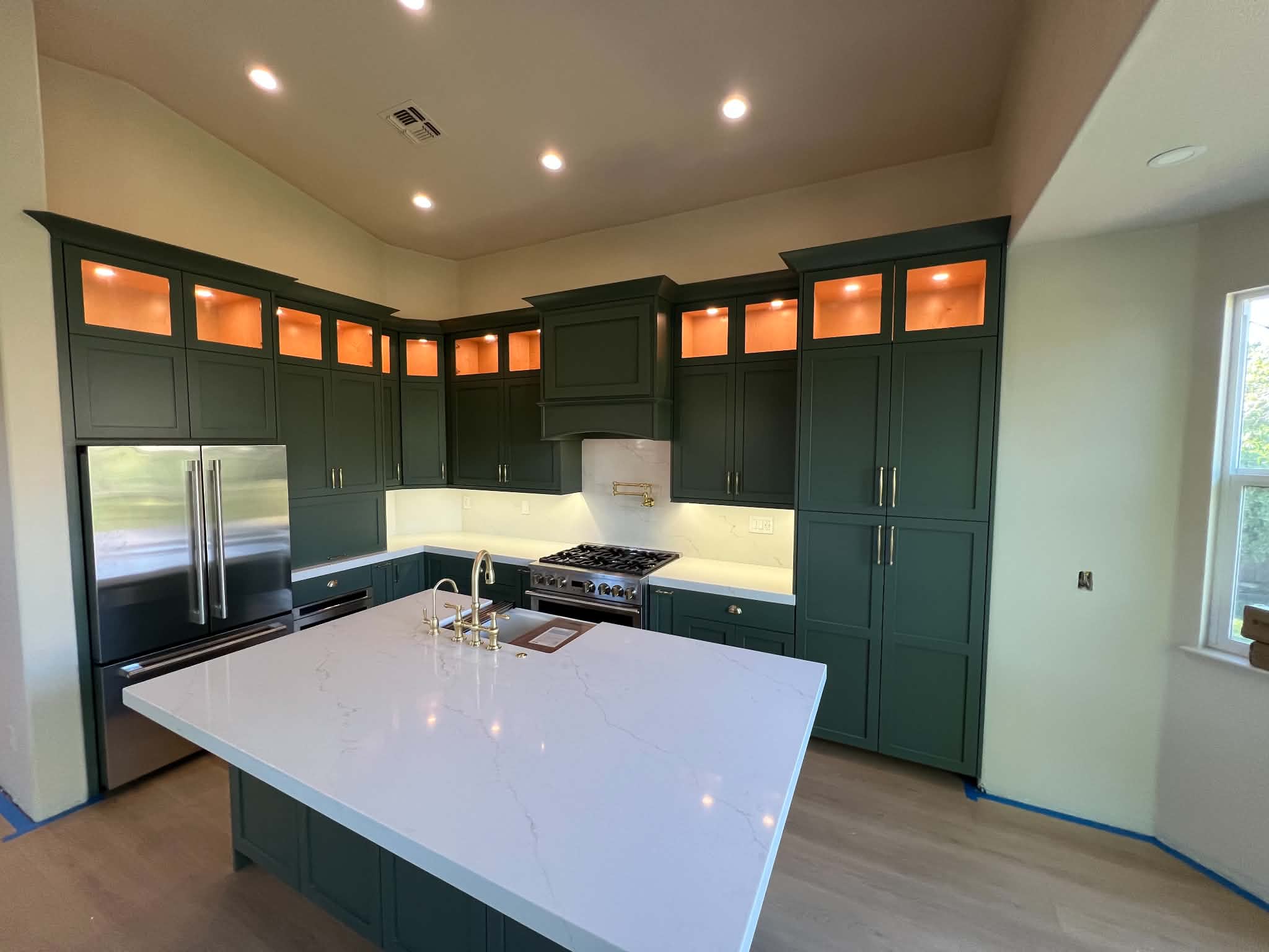 Deep green custom kitchen cabinetry with lit upper cabinets, brass hardware, and a white island countertop